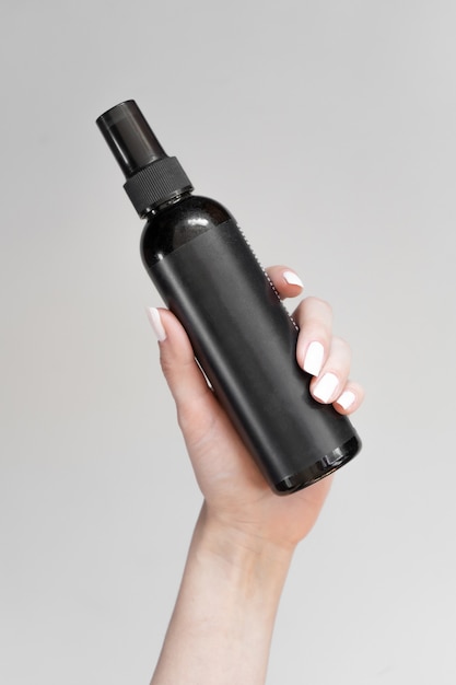 Una botella negra de spray cosmético con una etiqueta vacía en la mano de una mujer sobre un fondo claro