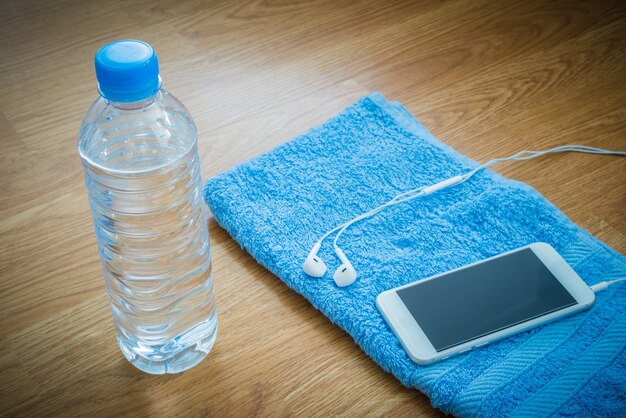 Botella plástica de agua, auriculares, teléfono inteligente y toalla en la mesa de madera