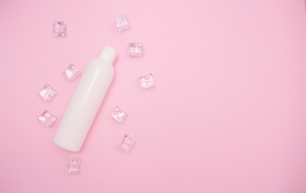 Botella de plástico blanco para champú u otros cosméticos sobre un fondo rosa entre cubitos de hielo