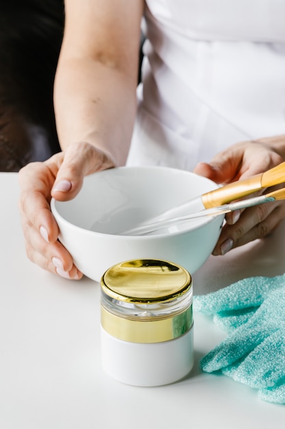 Botella de plástico marrón para cosméticos líquidos y cuenco de madera con palos de piedra para masaje facial en manos de mujer sobre mesa blanca.