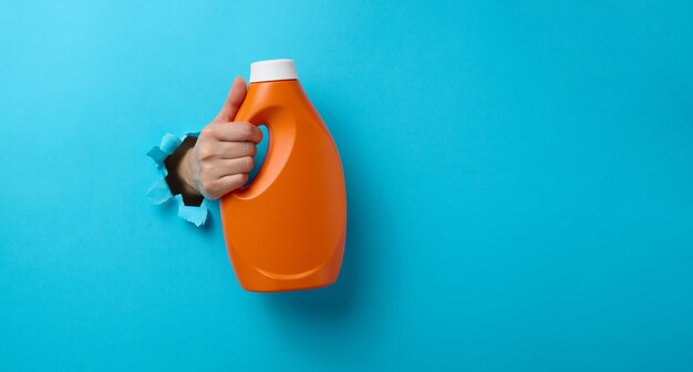 Botella de plástico naranja con detergente líquido en una mano femenina sobre un fondo rosa. Una parte del cuerpo sobresale de un agujero rasgado en el fondo, un lugar para una inscripción