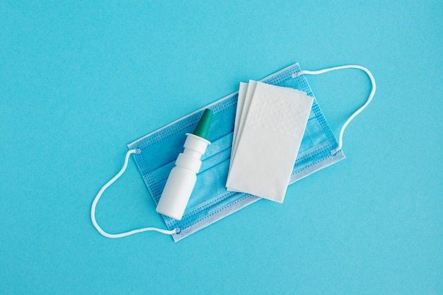 Botella de spray con gotas nasales, servilletas de papel y máscara médica sobre un fondo azul. El concepto de resfriado, gripe y secreción nasal.
