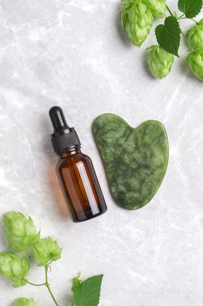 Botella de suero sin marca herramienta gua sha con lúpulo sobre fondo de mármol Jade gua sha para terapia de masaje facial Tratamiento antienvejecimiento y tonificante