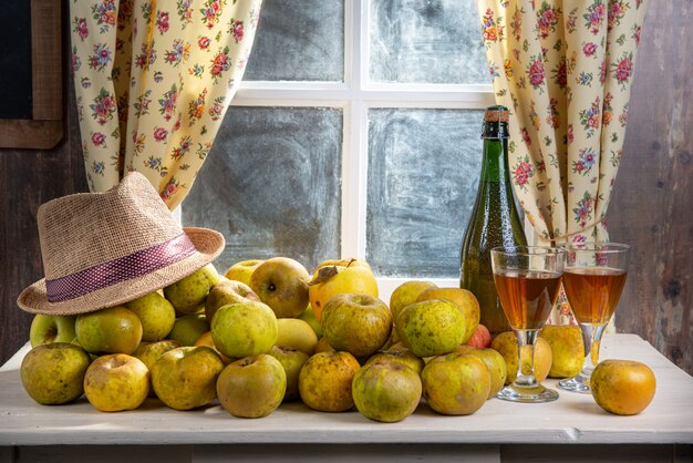 Botella y vasos de sidra con manzanas, cerca de la ventana, en casa rústica