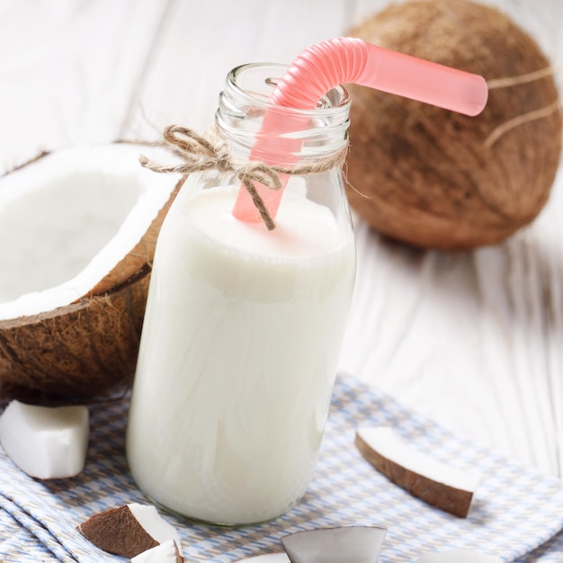 Botella de vidrio de leche o yogur en servilleta azul sobre mesa de madera blanca con coco a un lado