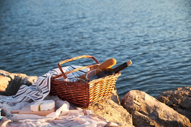Botella de vino con pan en una canasta de mimbre en una roca contra el mar durante la puesta de sol