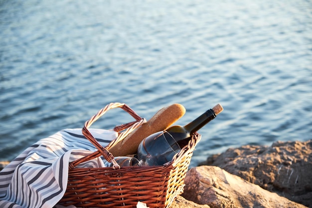 Botella de vino con pan en una canasta de mimbre en una roca contra el mar durante la puesta de sol