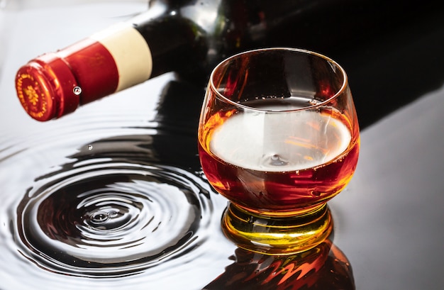 Botella de vino tinto con copa de vino. Salpicaduras de agua y gotas en el reflejo negro.