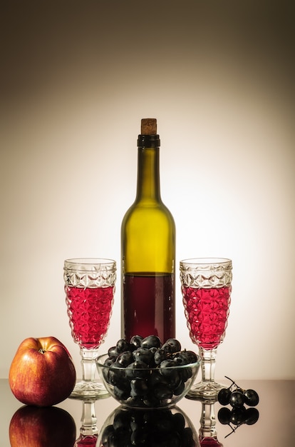 Botella de vino tinto, dos copas, una manzana y una copa con uvas de pie sobre una mesa