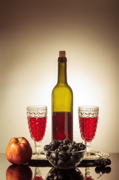 Botella de vino tinto, dos copas, una manzana y una copa con uvas de pie sobre una mesa