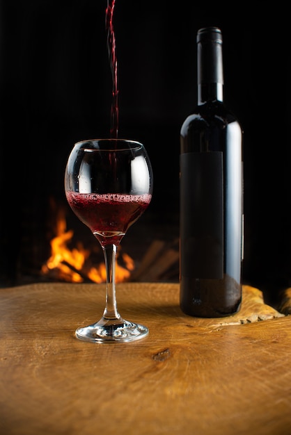 Botella de vino y vaso que se llena con vino sobre la superficie de madera rústica con fuego detrás