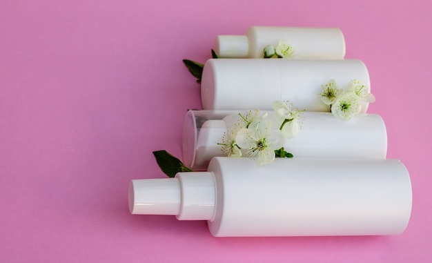Botellas de cosmética blanca, bomba de baño, jabón artesanal, sal de baño, cepillo de masaje, esponja, bastoncillos de algodón con flores de cerezo sobre un fondo rosa. Concepto de cosmética orgánica natural.