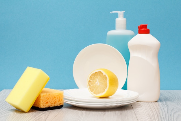Foto botellas de plástico de líquido para lavar platos, limpiador de vidrio y azulejos, platos limpios, limón y esponjas sobre fondo azul. concepto de lavado y limpieza.