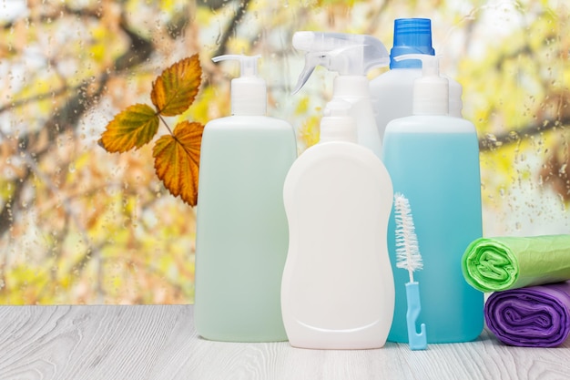 Foto botellas de plástico de líquido para lavar platos, limpiador de vidrios y azulejos, detergente para hornos microondas y estufas y bolsas de basura frente a la ventana con gotas de agua y hojas de otoño.