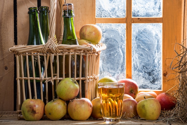 Botellas y vaso de sidra con manzanas cerca de la ventana