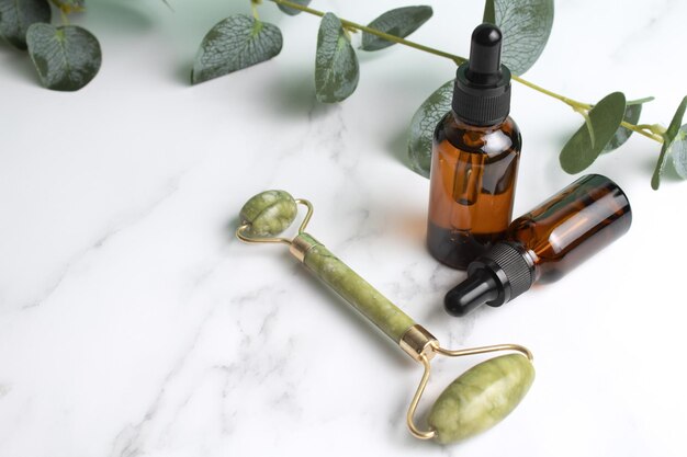 Botellas de vidrio con rodillo facial de jade de aceite para masaje facial sobre fondo de mármol con hojas de eucalipto Cuidado cosmético de la piel facial y spa Concepto de tratamiento natural