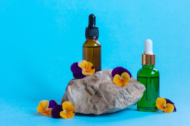 Botellas de vidrio de suero con pipeta y hermosas flores de viola sobre fondo azul. Concepto cosmético de spa orgánico natural. Vista frontal.