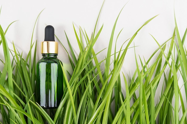 Botellas de vidrio verde con suero, aceite esencial, colágeno u otro producto cosmético entre la hierba verde sobre fondo blanco. Vista superior de la maqueta del concepto cosmético de spa orgánico natural.