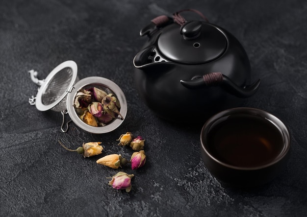 Botões de rosa misturam chá no infusor de filtro e bule de cerâmica e xícara em fundo escuro