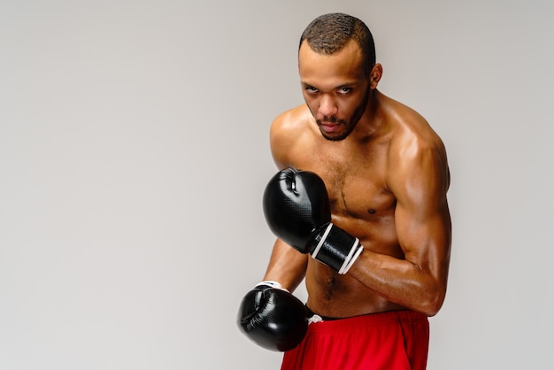 Boxeador africano joven confiado en guantes de boxeo que se colocan sobre la pared gris clara