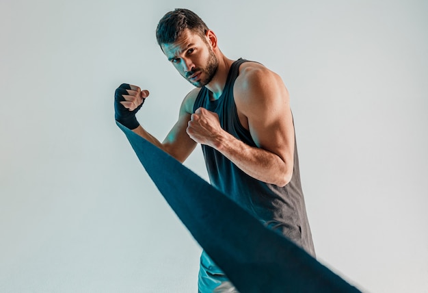 Boxeador concentrado con vendaje de boxeo en mano. Joven deportista europeo barbudo con uniforme deportivo y mirando a cámara. Aislado sobre fondo gris con luz turquesa. Sesión de estudio