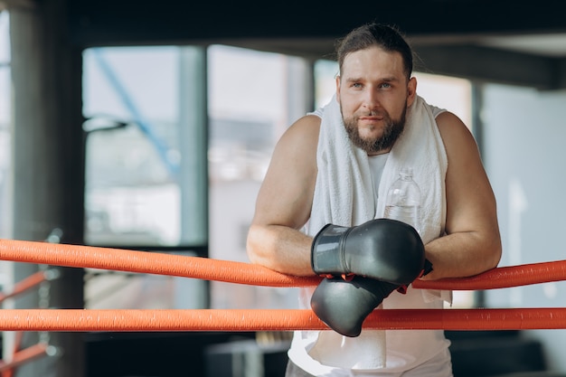 Boxeador deportivo sudoroso con vendas de boxeo en las manos mirando a la cámara desde el ring mientras toma un descanso después de hacer ejercicio en el gimnasio