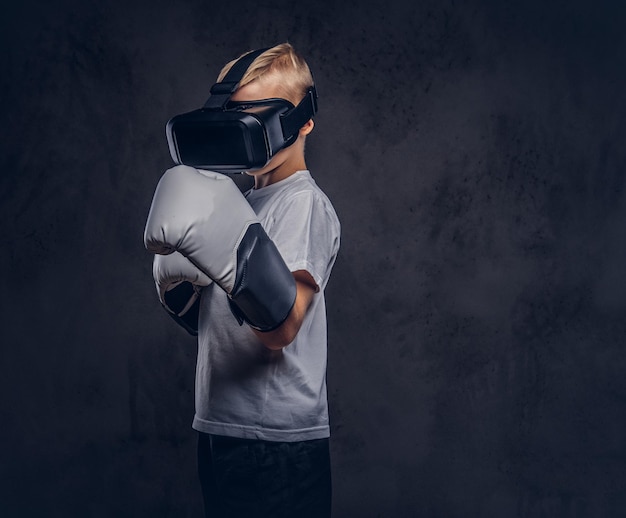 Boxeador escolar con cabello rubio vestido con una camiseta blanca con gafas de realidad visual y guantes de boxeo, entrena en un estudio. Aislado en un fondo de textura oscura.