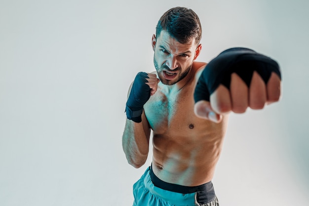 El boxeador europeo barbudo joven enojado está boxeando. El deportista usa vendas de boxeo en los brazos y mirando a la cámara. Aislado sobre fondo gris con luz turquesa. Sesión de estudio. Copia espacio