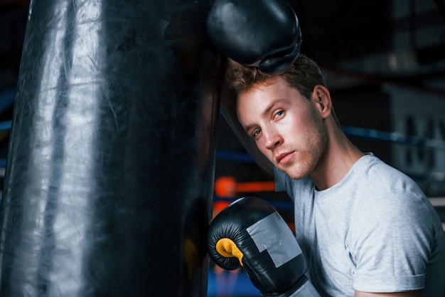 Boxeador joven cansado con camisa blanca y guantes protectores apoyado en saco de boxeo en el gimnasio.