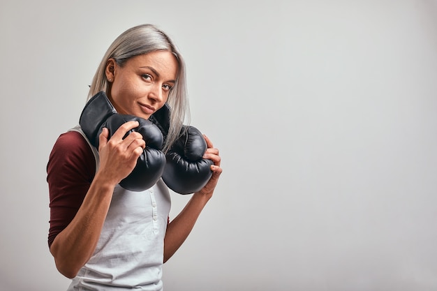 Boxeador joven hermosa mujer sexy posando con guantes de boxeo negros en sus manos sobre un fondo gris. Copie el espacio, fondo gris, logro de objetivos.