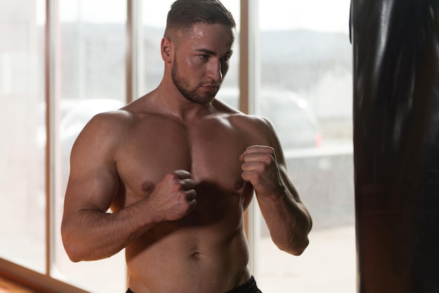 Foto boxeador muscular descamisado con el saco de arena en gimnasio