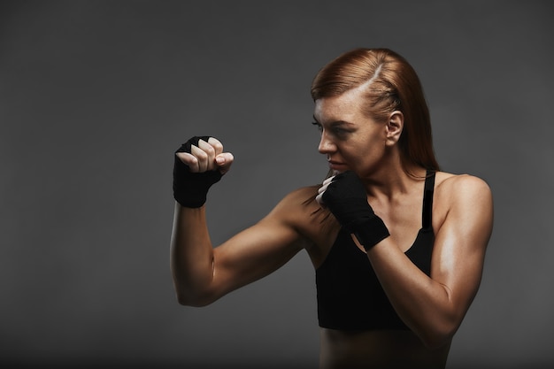 Boxer feminina com as mãos em bandagens pretas em posição de boxeador em uma superfície cinza escura, posando com um pedido de proteção