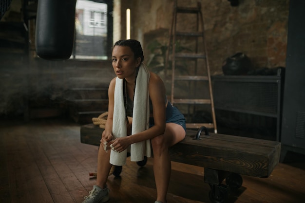 Boxer mulher relaxada descansando no banco com a toalha no pescoço. Menina cansada em roupas esportivas sentada sobre o velho interior sujo do ginásio