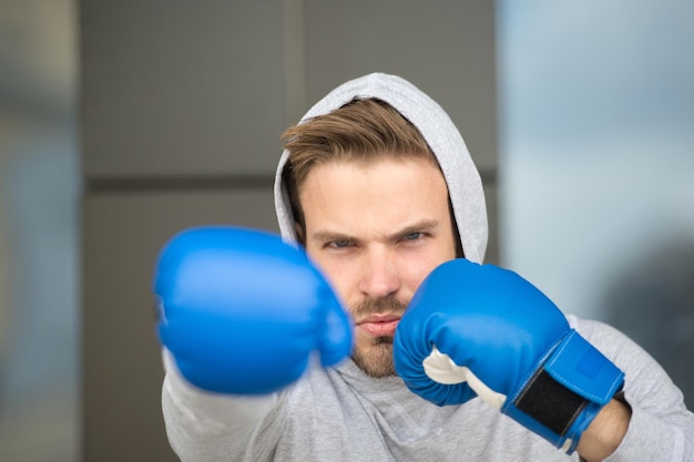 Boxkonzept. Mann Athlet auf konzentriertem Gesicht mit Sporthandschuhen, die Boxen üben, urbaner Hintergrund. Boxer mit Kapuze auf dem Kopf übt Jab-Punch. Sportlerboxertraining mit Boxhandschuhen.