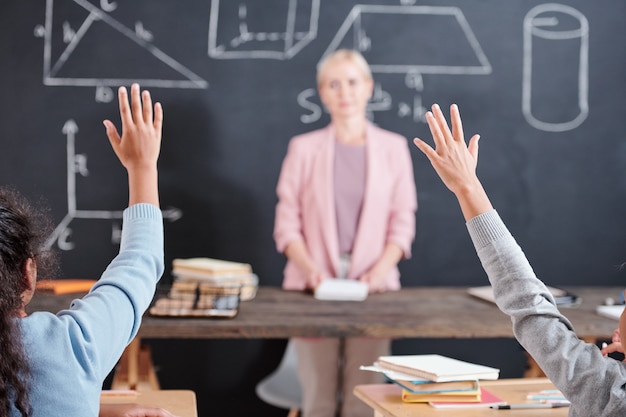 Braços de duas crianças de escola inteligentes dispostas a responder à pergunta do professor em pé no quadro-negro com formas geométricas na aula