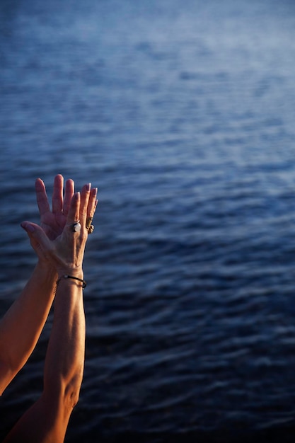 Braços de uma mulher realizando um mudra com o fundo do mar ao pôr do sol