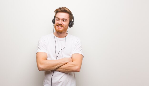 Braços seguros e de cruzamento do homem novo do ruivo que olham acima, olhando acima. Ouvir música com fones de ouvido.