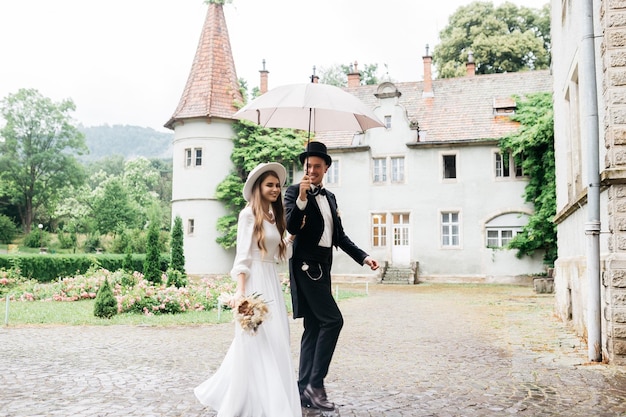 Bräute in Hüten Junges Mädchen in einem weißen Hochzeitskleid und Hut mit einem Blumenstrauß Die Braut und der Bräutigam gehen und halten einen Regenschirm Bräute auf dem Hintergrund des Schlosses