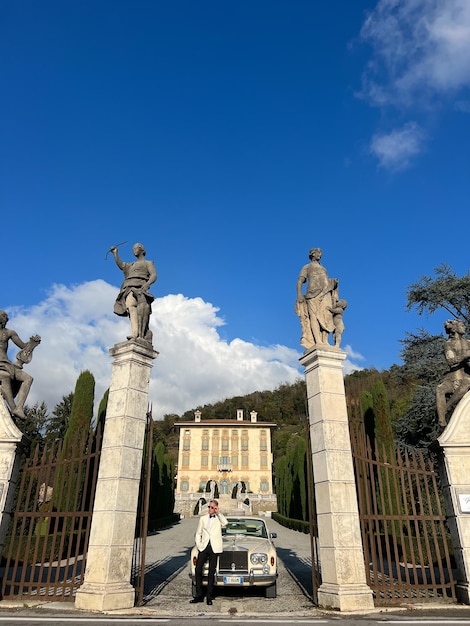 Bräutigam mit Sonnenbrille steht neben dem Auto am Tor der Villa mit Skulpturen auf Sockeln