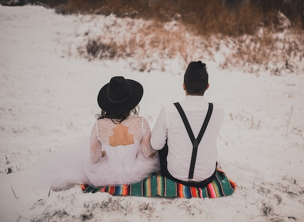 Bräutigam sitzt mit Braut im weißen Hochzeitskleid und im schwarzen Hut auf mexikanischem Nationalschleier auf schneebedecktem Hügel