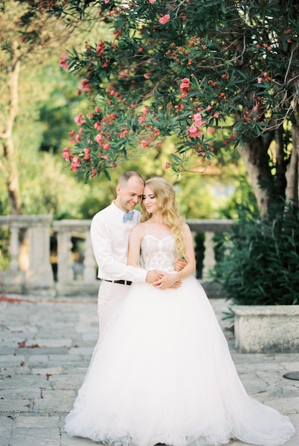Bräutigam umarmt Braut, die unter einem Baum mit rosa Blumen im Garten steht