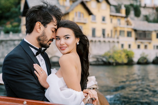 Bräutigam umarmt die Braut, berührt die Stirn, steht auf einer Yacht, die auf dem Lake Como in Italien segelt
