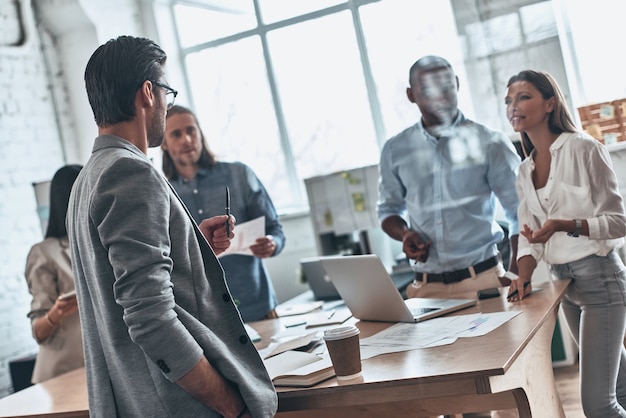 Brainstorming-Meeting. Gruppe junger moderner Menschen, die miteinander kommunizieren, während sie hinter der Glaswand im Sitzungssaal arbeiten working