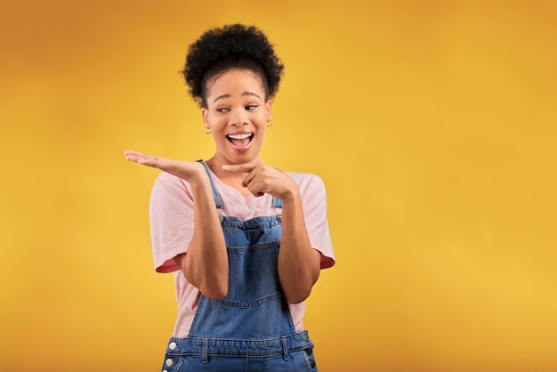 Branding-Werbung und eine schwarze Frau, die auf ihre Handfläche zeigt, um ein Produkt auf gelbem Hintergrund im Studio zu bewerben. Smile-Marketing oder Space mit einer glücklichen jungen Markenbotschafterin