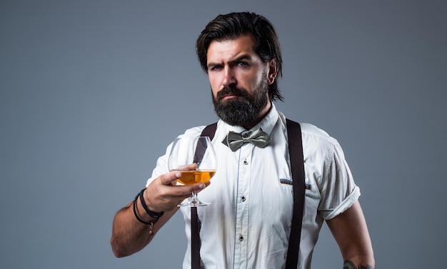 Brandy o coñac hombre barbudo en tirantes bebe whisky escocés tipo brutal barman usa pajarita elegante barman masculino guapo hipster bebiendo ron vidrio fiestero con alcohol