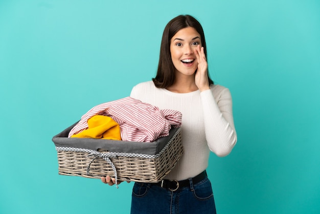 Brasileira adolescente segurando uma cesta de roupas isolada em um fundo azul gritando com a boca aberta