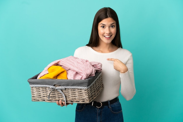 Brasileira adolescente segurando uma cesta de roupas isolada em uma parede azul com expressão facial surpresa