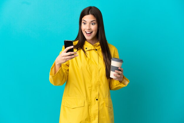 Brasileira adolescente vestindo um casaco à prova de chuva sobre um fundo azul isolado segurando um café para levar e um celular