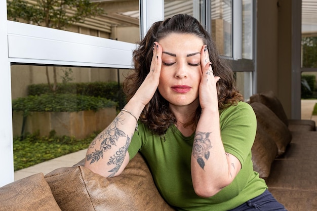 Brasileira tatuada e preocupada com as duas mãos na testa dela sentada no sofá