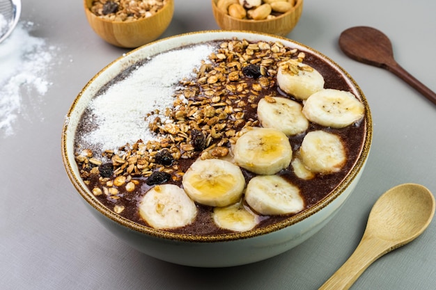 Brasilianische Acai in einer weißen Schüssel mit Banane, Milchpulver, Honig, Müsli und Nüssen in kleinen Bambusschalen Cashewnüsse und Para Grauer Hintergrund Selektiver Fokus